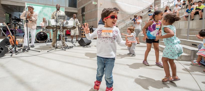 Young child with sunglasses dancing with a band playing behind them. Other children dancing about int he background in amphitheater setting.