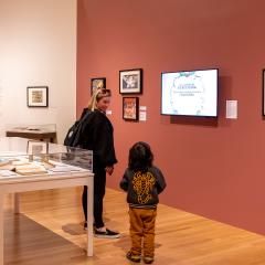 An adult and child look at a screen on a red gallery wall. Around them are framed artworks, tables displaying open books, and a large illustration of a monster seeming to hang from the top of a wall.